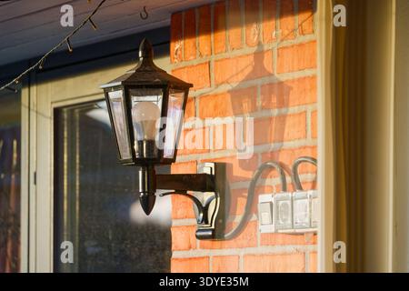 A classic black metal outdoor wall lantern with a light bulb mounted on a red brick wall illuminated by warm sunlight casting shadows. Stock Photo