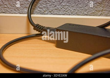 Black power adapter with connected cable resting on a wooden surface against a textured wall background, showing electronic device charging equipment. Stock Photo