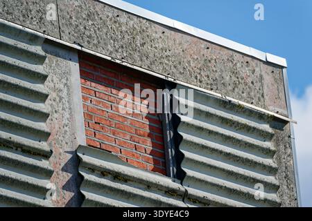 Weathered industrial wall featuring corrugated metal panels partially removed to reveal red brickwork beneath against a clear blue sky background. Stock Photo
