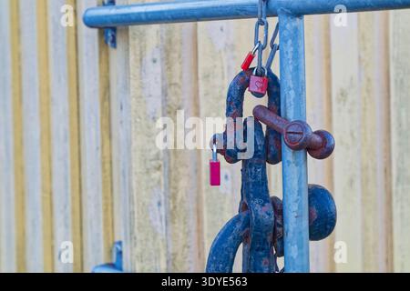 A close view of a rusty metal chain locked with red padlocks on a blue metal gate against a weathered wooden wall background. Stock Photo