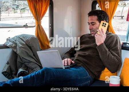 Man talking on mobile phone and using laptop inside a parked camper van. Digital nomad lifestyle, freedom and travel Stock Photo