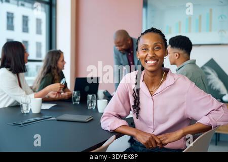 Young businesswoman smiling confidently at camera while diverse team discusses project in boardroom Stock Photo