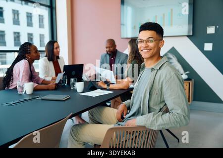 Young businessman in smart casual smiling at camera while diverse team collaborates in boardroom at office Stock Photo