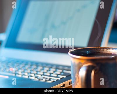 Laptop screen shows upward-trending graph. Coffee mug steams beside keyboard. Work session suggests focus and productivity. Soft lighting enhances cal Stock Photo