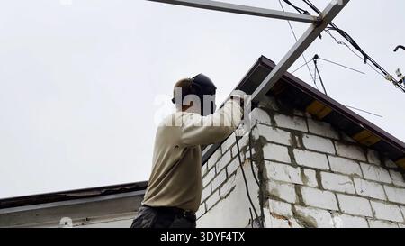 Manual laborer in protective mask welds iron detail at construction. Professional young repairman with welding machine working at the site. Male Stock Photo