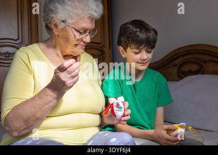 Grandmother and grandson making handmade Easter decorations together at home. Elderly woman sewing ribbon on decorative Easter eggs while spending tim Stock Photo