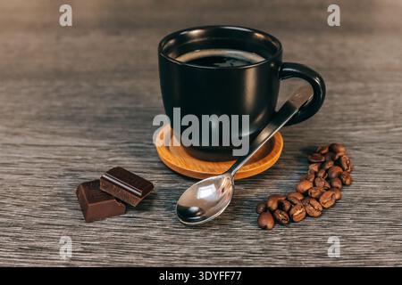 Black cup of coffee filled with dark coffee with coffee beans, a spoon and 2 dark chocolates, everything on a grey table - Cozy breakfast scene Stock Photo