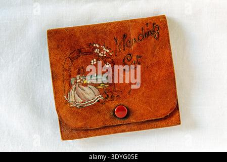 Vintage Leather Mending Set or Sewing needle Case with girl or woman in long white dress sitting sewing on bench with seascape behind Stock Photo