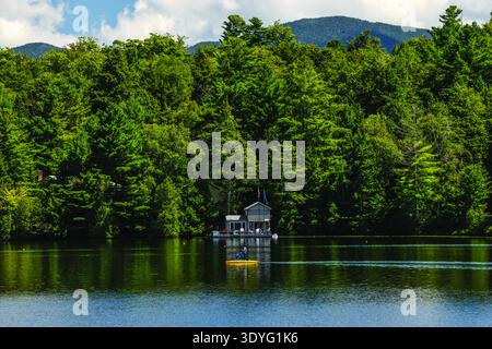 A lone person paddleboarding on a calm lake beside a small cabin in dense green forest beneath distant mountains, conveying a peaceful summer day. Stock Photo