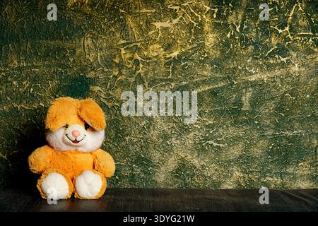 A soft orange stuffed bunny sits on a dark surface in front of a green wall with a rough texture. Stock Photo