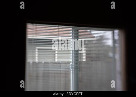 Blinds half-closed frame a quiet, muted street view. Soft light filters through slats, creating gentle shadows. Reflections hint at a calm, unremarkab Stock Photo