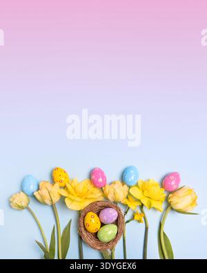 Easter colored painted eggs in bird nest with yellow daffodils, white tulips on blue pink gradient background with space for text and greetings. Easter and spring concept. Stock Photo