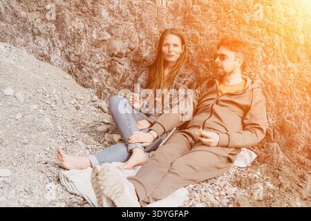 Couple beach relaxation young man and woman sitting on pebble beach against rocky cliff sharing snacks in golden sunlight Stock Photo