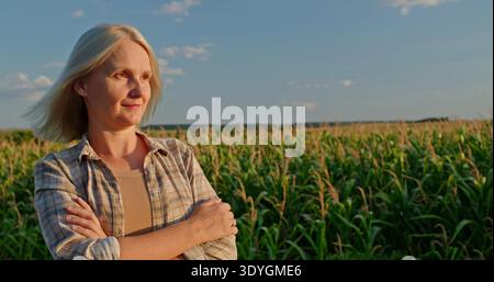 Confident woman standing in a cornfield at sunset, wearing a plaid shirt, arms crossed, with a serene expression. Stock Photo