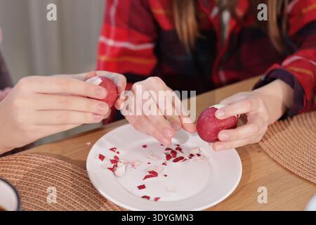 Two people peeling red dyed Easter eggs over a white plate, with broken eggshell pieces scattered across the surface. Stock Photo