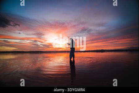 Man standing barefoot in shallow water at sunset, operating a drone controller Stock Photo