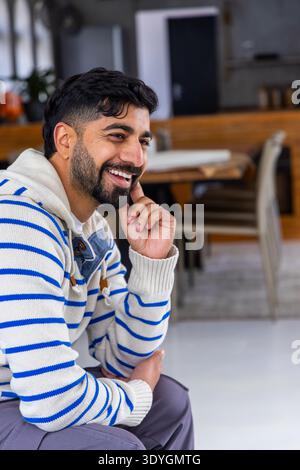 Indian man sitting in cafe, smiling and supporting chin, wearing striped hooded sweater, copy space Stock Photo
