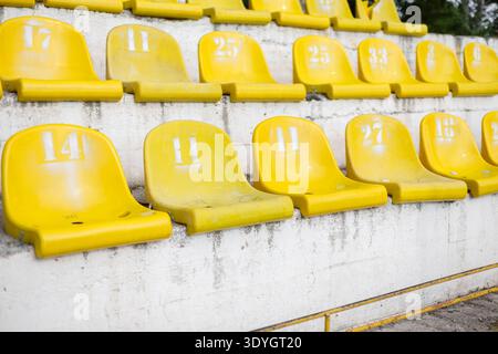 Rows of weathered yellow stadium seats with white painted numbers on a concrete grandstand. Close-up of numbered yellow spectator seating at an old ou Stock Photo