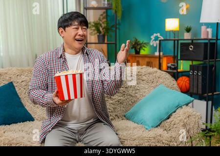 Asian man laughs loudly watching comedy movie, holding stomach and eating popcorn, sitting on home sofa. Chinese guy enjoys humorous entertainment, relaxed joyful reaction fills room with carefree fun Stock Photo