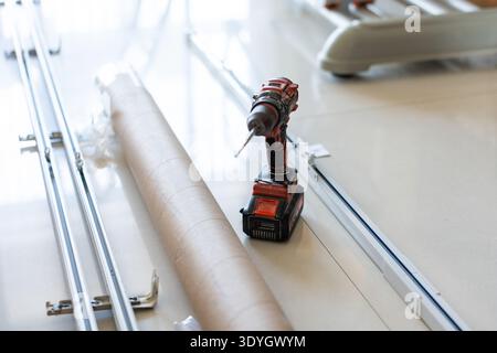 Cordless drill with screwdriver bit resting on floor near metal rods and cardboard tube in indoor Stock Photo