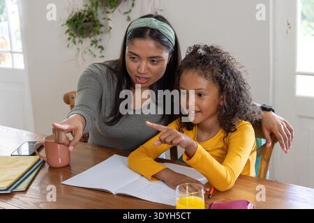 Asian woman leaning in, pointing with child at wooden table over open notebook and pencil Stock Photo