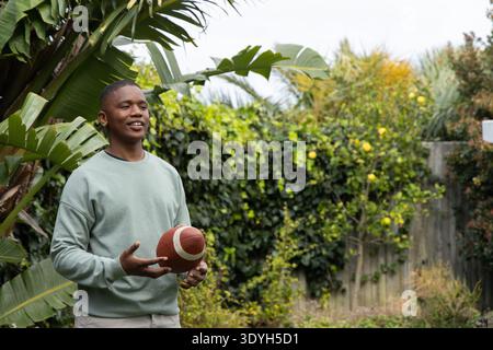 African American man standing in backyard garden holding American football among banana leaves Stock Photo