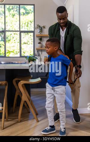 African American father and son helping child put on backpack in home kitchen near island Stock Photo