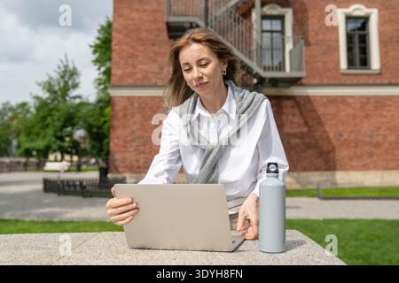 Cheerful businesswoman working on laptop outside in park, preparing for online business conference. Stock Photo