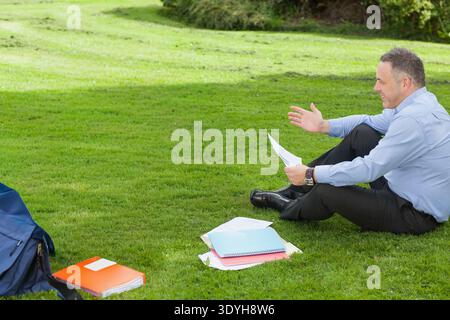 Man sitting on lawn wearing light blue shirt and dark pants, holding papers, backpack, copy space. Outdoor, executive, discussion, documents, notebook Stock Photo