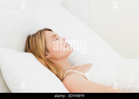 Mid adult female wearing white camisole reclining on bed, pillow against textured white headboard. Minimal, serene, tranquil, calm, diffuse, soft, lin Stock Photo