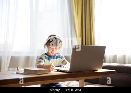 child boy in earphones is using a laptop and study online at home. homeschooling, distant learning || Model released Stock Photo