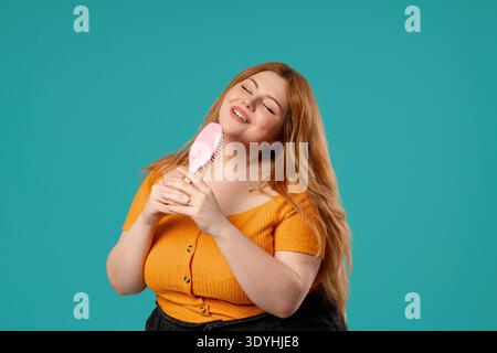 Body positive woman Singing, Dancing With Hair Brush Instead Microphone On Blue. Curvy Lady having fun, listening to music, karaoke, dreams of being celebrity. High quality || Model released Stock Photo