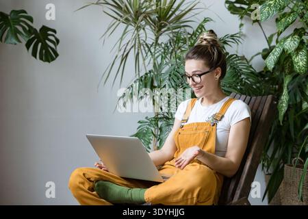 Woman gardener in glasses wearing overalls, sitting on wooden chair in greenhouse, using laptop after work, talks with friend on video call. Working home. || Model released Stock Photo