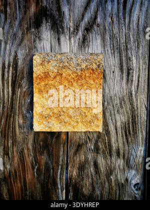 A square, rough textured material, changing from grey to golden-orange, is secured by two screws onto a surface of dark, Stock Photo