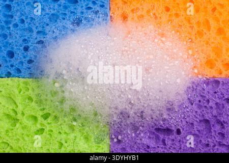 Close-up of four brightly colored household sponges arranged in a grid formation with a mound of soap bubbles in the center. Vivid colors - blue, oran Stock Photo
