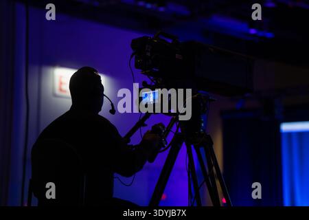 Silhouette of a broadcast camera operator monitoring a live stage performance on a professional video rig during a corporate media event in a blue lit Stock Photo
