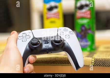 Hand holding a white gaming controller with snacks in the background. Stock Photo