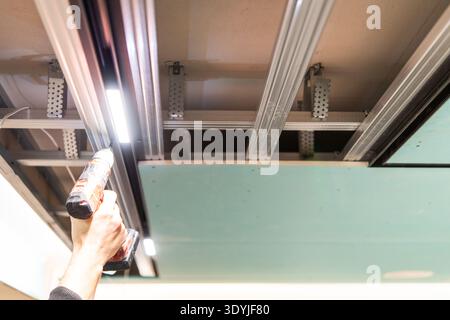 Hand using cordless drill to install LED track lighting on the metal framework of a suspended ceiling in a modern interior space. Stock Photo