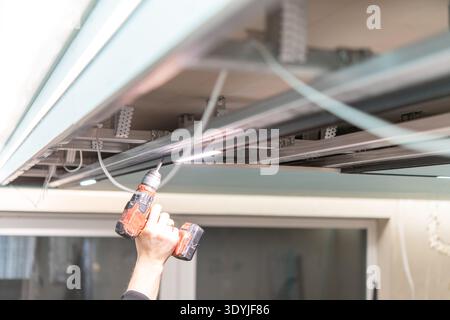 Hand with power drill installs LED track lighting on suspended ceiling framework in modern interior under construction. Stock Photo