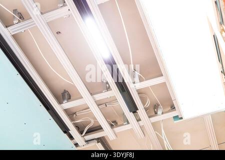 Close-up of a modern suspended ceiling installation featuring LED track lighting, exposed white wiring, and metallic support beams indoors. Stock Photo