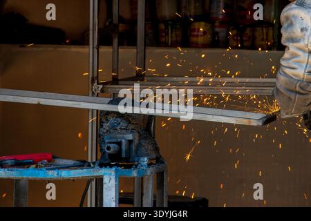 A worker  uses a grinder on a metal frame, creating a spray of glowing orange sparks in a workshop. Stock Photo