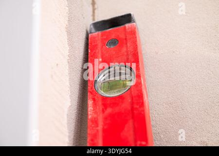 Close-up of a red spirit level tool measuring the vertical alignment of a beige textured wall surface. Stock Photo