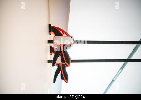 Closeup of red and black clamps securing wooden boards against a wall during carpenter furniture assembly. Stock Photo