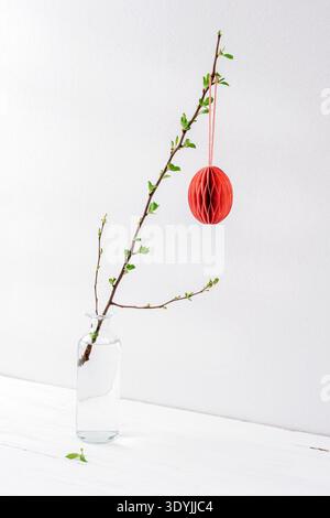 Easter decoration and spring twig in glass vase on white wooden table against white wall. Closeup. Stock Photo