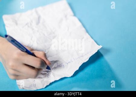 front view of hand with pen writing on crumpled sheet of paper close up on blue paper background Stock Photo