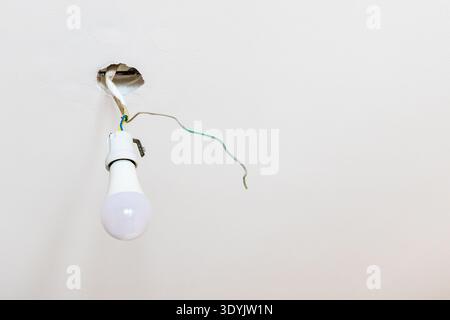 LED light bulb in electrical socket hangs on electrical wires from hole in ceiling during apartment renovation Stock Photo