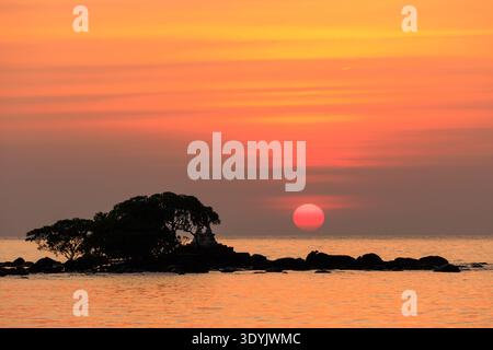 Sun setting behind a small island with trees and rocks, reflecting orange light on ocean water Stock Photo
