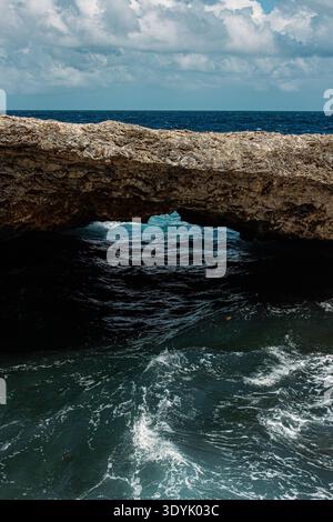 An ancient natural bridge over a deep blue ocean, with wild water and blue skies. Stock Photo