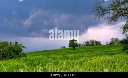 Stormy sky over green hills in Neviano degli Arduini, Italy.A dramatic landscape featuring vibrant green spring meadows under a dark, heavy storm sky. Stock Photo