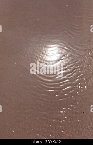 Close-up view of water flowing over wet sand, creating dynamic, natural patterns, featuring ripples and reflections of sunlight on the shallow water, Stock Photo